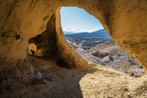 View from inside a cave, showcasing a bright landscape with mountains and blue sky in the background.