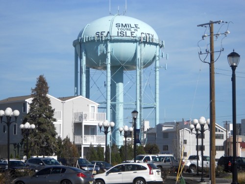 A blue water tower with the text "SMILE, YOU'RE IN SEA ISLE CITY" surrounded by buildings and parked cars.