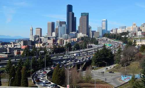 A bustling highway with traffic, leading to a skyline of tall buildings in a city, surrounded by greenery.