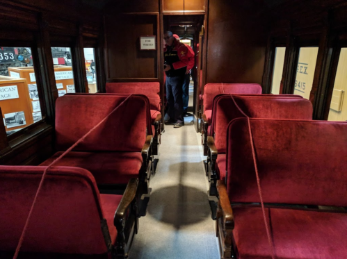 Interior of a vintage train car with red velvet seats and a person standing in the aisle.
