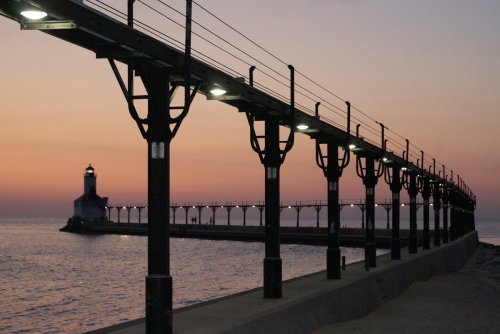 A pier extends into the water at sunset, with a lighthouse in the distance and lights illuminating the walkway.