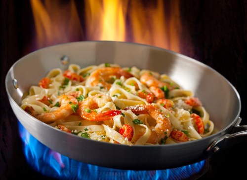 A skillet of shrimp fettuccine pasta with herbs, set against a backdrop of flames.