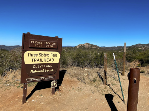 Sign for Three Sisters Falls Trailhead in Cleveland National Forest, with a clear blue sky and mountainous background.