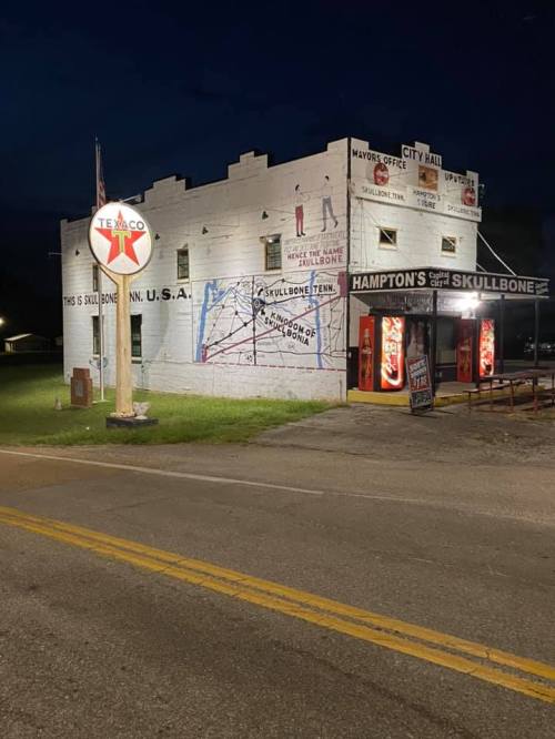 A vintage building at night with signs for "Skullbone" and "Hampton's," featuring a Texaco gas station logo.
