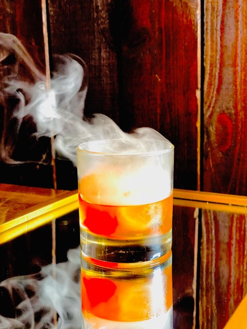 A glass of cocktail with smoke rising, reflecting on a shiny surface against a wooden background.