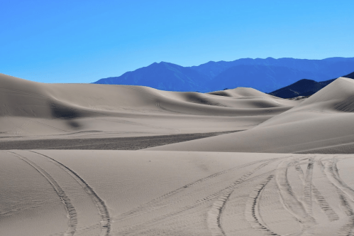 A vast desert landscape with rolling sand dunes and distant mountains under a clear blue sky.