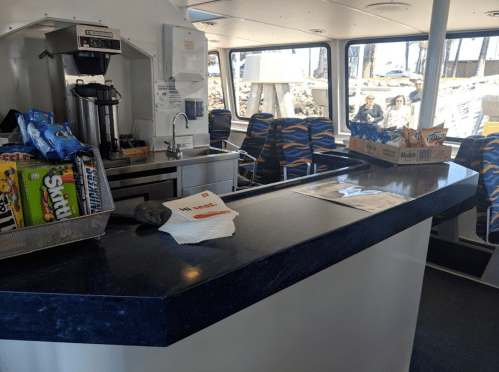 A boat interior featuring a snack bar with drinks, snacks, and seating, with people visible outside the window.