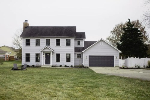 A two-story gray house with a garage, surrounded by a lawn and a white fence, on a cloudy day.