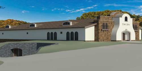 A modern spa building with a stone facade, arched windows, and a sloped landscape under a blue sky.
