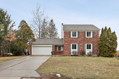 A two-story brick house with a garage, surrounded by trees and a grassy yard on a clear day.