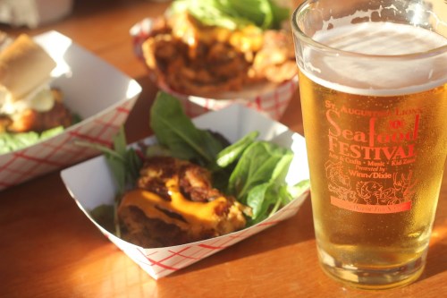 A glass of beer next to seafood dishes served in paper trays, with lettuce and a festive logo on the glass.