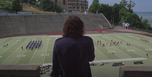 A person stands on a stadium balcony, overlooking a football field where groups are practicing drills.