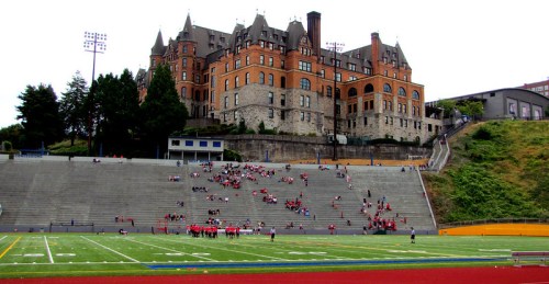 A historic building overlooks a football field with spectators and players on a cloudy day.