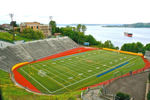 A panoramic view of a sports field with bleachers, surrounded by greenery and a body of water in the background.