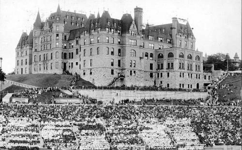Historic black-and-white photo of a large crowd in front of a grand castle-like building on a hillside.