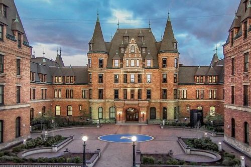 Historic brick building with turrets, surrounded by a courtyard and landscaped gardens under a cloudy sky.