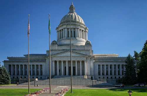 The Washington State Capitol building with a large dome, flanked by flags and surrounded by greenery and flowers.