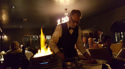 A chef in a tuxedo prepares a flaming dish at an elegant restaurant, with diners enjoying their meals in the background.