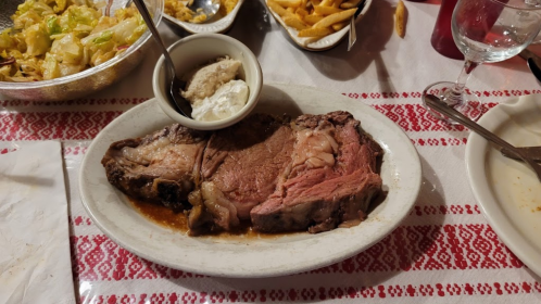A large serving of prime rib with a side of creamy horseradish sauce, accompanied by fries and a salad in the background.