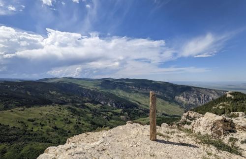A wooden post stands on a rocky outcrop, overlooking a vast green valley and distant mountains under a blue sky.