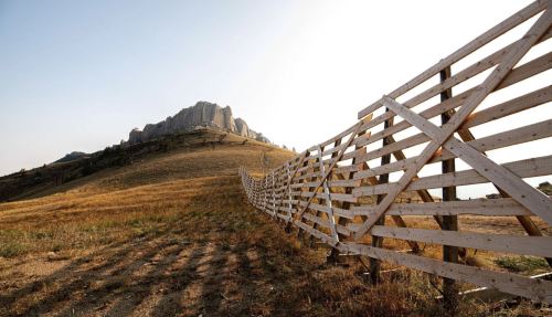 A wooden fence stretches across a grassy landscape, leading towards a rocky hill under a clear sky.