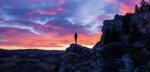 A silhouette of a person standing on a rocky outcrop against a vibrant sunset sky with colorful clouds.