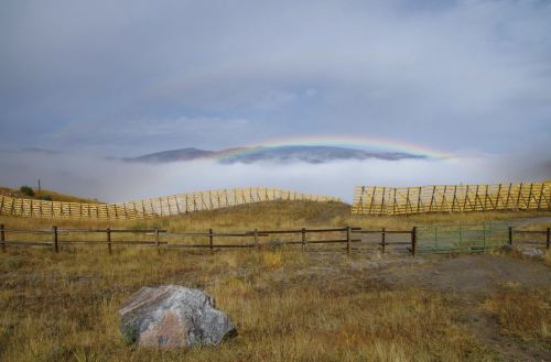 A wooden fence curves across a grassy landscape under a cloudy sky with a rainbow arching above.
