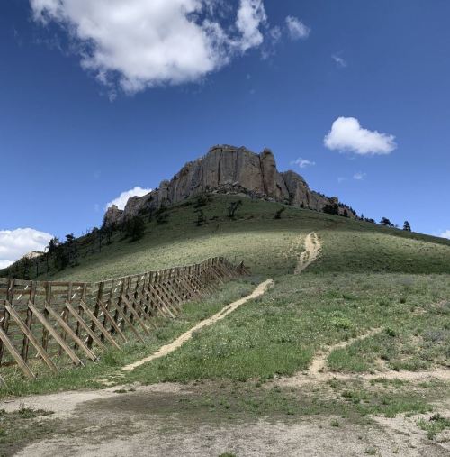 A rocky hill rises against a blue sky with clouds, surrounded by green grass and a wooden fence in the foreground.