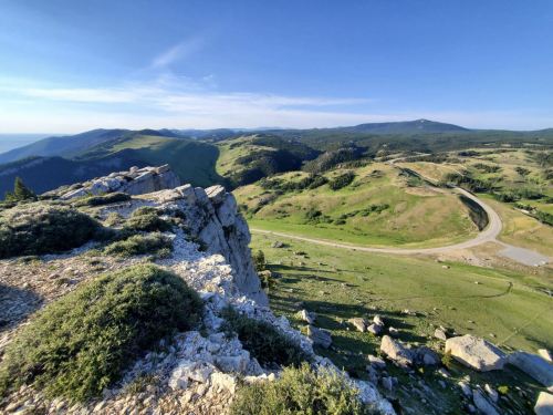 A panoramic view of rolling green hills and rocky cliffs under a clear blue sky. A winding road is visible in the distance.