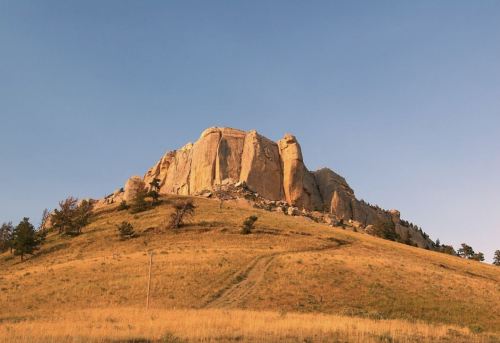 A rocky cliff rises above a grassy hillside under a clear blue sky, with sparse trees dotting the landscape.