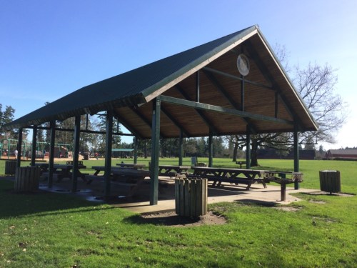 A large, open picnic shelter with a green roof, surrounded by grass and trees in a sunny park setting.