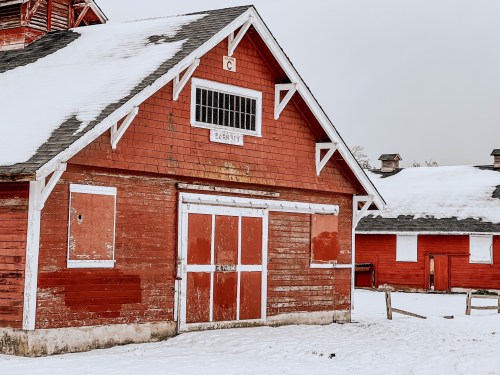 A weathered red barn with white trim, surrounded by snow, under a gray sky. Nearby buildings are also visible.
