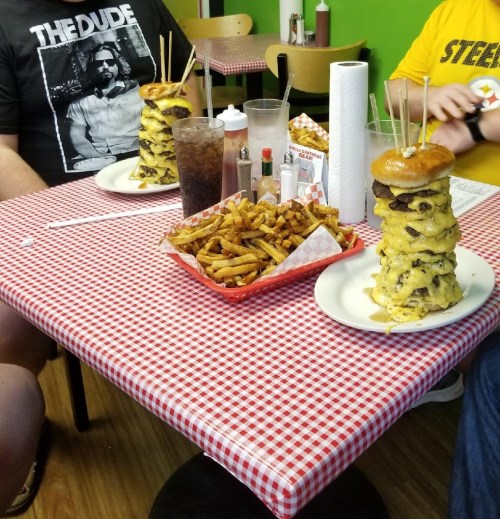 A table with a large stacked burger, fries, and drinks, with diners enjoying a casual meal in a checkered setting.