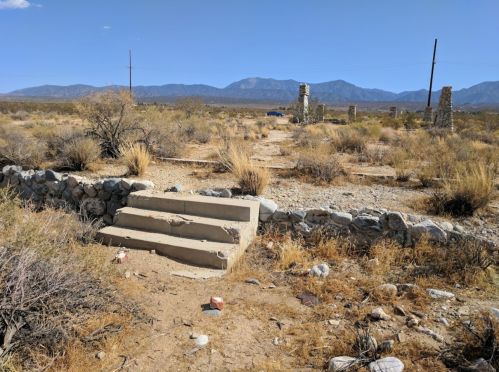 Desert landscape with stone steps leading to ruins, surrounded by dry vegetation and distant mountains under a clear blue sky.