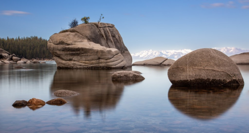 Serene lake scene with smooth rocks and a distant mountain range under a clear blue sky.