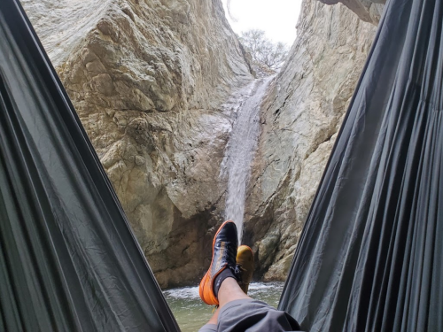 A person relaxes in a hammock with a waterfall and rocky cliffs in the background.