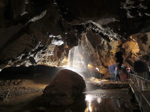 A dimly lit cave with a waterfall, large rock formations, and visitors exploring the area.
