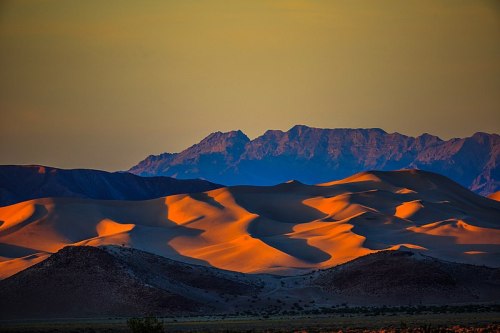 Golden sand dunes contrast with distant mountains under a colorful sunset sky.