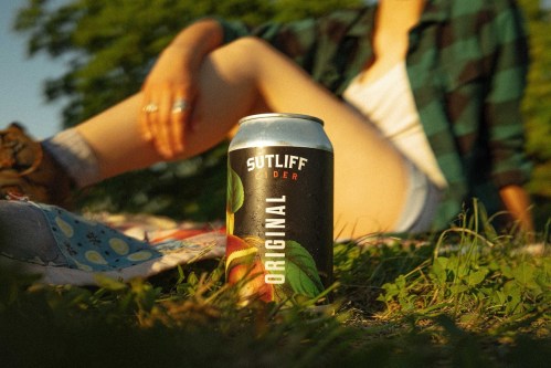 A can of Sutliff Original cider sits on grass, with a person lounging in the background on a sunny day.
