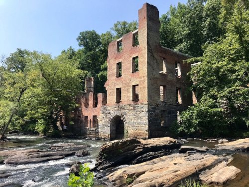 Ruins of a brick building beside a river, surrounded by lush greenery and rocky terrain under a clear blue sky.