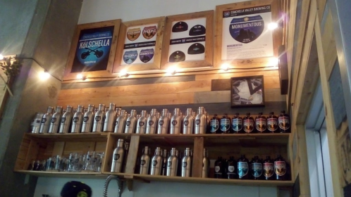 A wooden shelf displaying various beer bottles and growlers, with framed awards and posters above. Warm lighting enhances the ambiance.