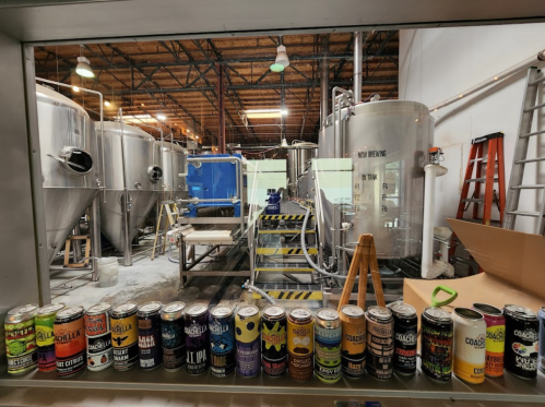 A brewery interior featuring large fermentation tanks and a display of various colorful beer cans in the foreground.