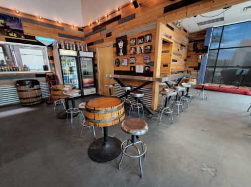 A cozy bar area with wooden walls, barrel tables, and stools, featuring a fridge and artwork on the walls.