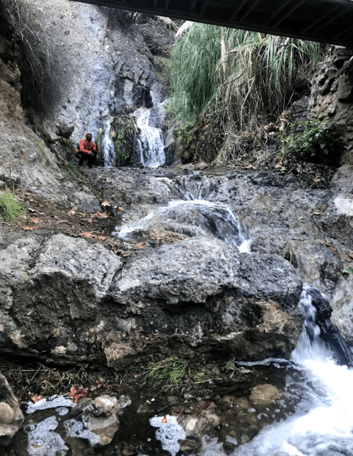 A person in an orange jacket sits near a rocky stream with small waterfalls, surrounded by greenery and rugged terrain.