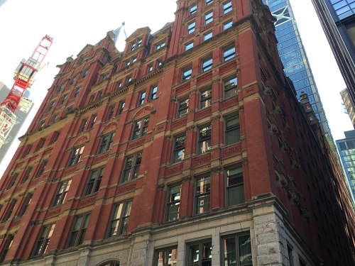 Historic red brick building with ornate details, surrounded by modern skyscrapers in an urban setting.
