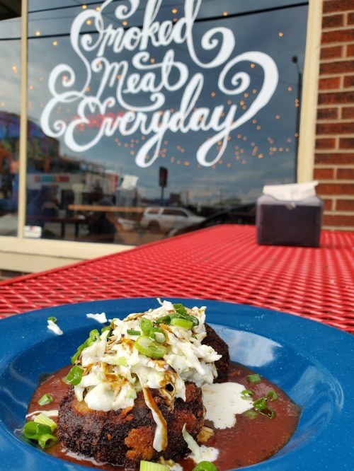 A plate of smoked meat topped with coleslaw and green onions, set against a restaurant window with decorative text.