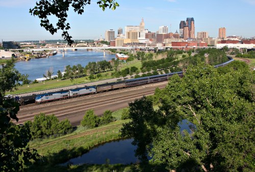 A train travels along tracks near a river, with a city skyline and green trees in the background on a clear day.