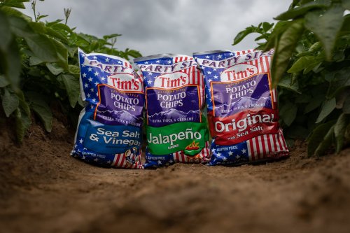 Three bags of Tim's Potato Chips in a field: Sea Salt & Vinegar, Jalapeño, and Original, with a cloudy sky above.