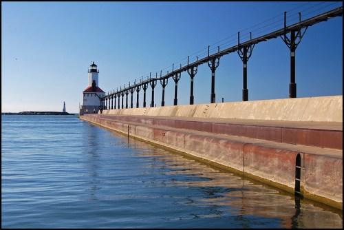 A lighthouse stands at the end of a long pier, reflecting in calm waters under a clear blue sky.