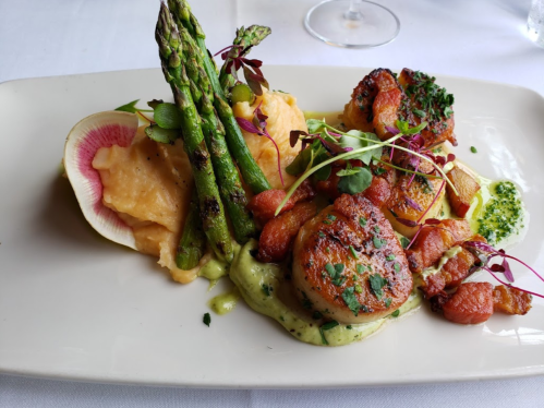 A gourmet dish featuring scallops, asparagus, sweet potato puree, and vibrant garnishes on a white plate.